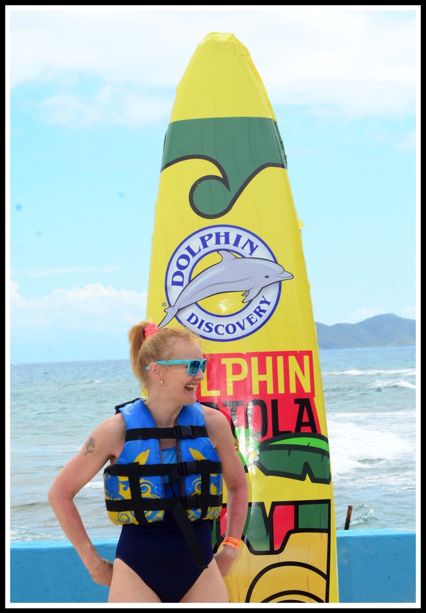 Sarah wearing swim suit and life jscket, stood in front of a large surf board promoting the dolphins