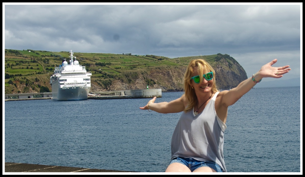 Sarah is sat on a wall on the right with her arms stretched out while the cruise ship is facing us on the left 