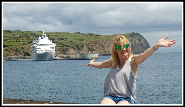 Sarah is sat on a wall on the right with her arms stretched out while the cruise ship is facing us on the left 