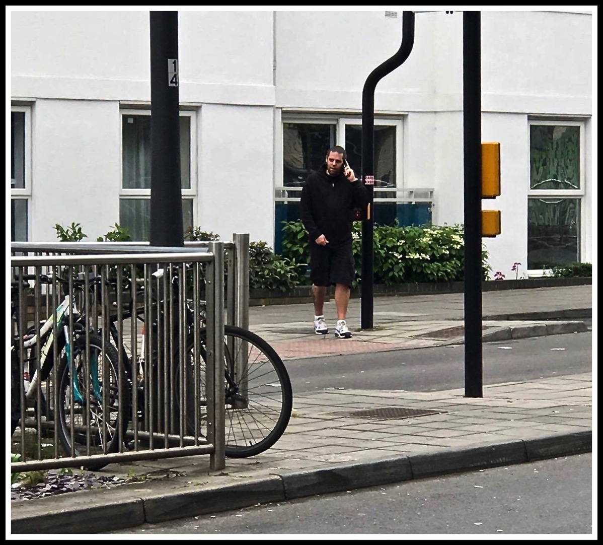 A photo of me crossing road to train station while talking on the phone wearing a black hoody, black shorts and black and white trainers
