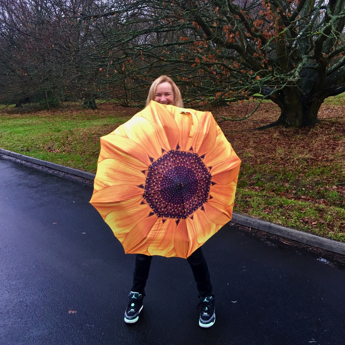 Sarah walking along a wet road with a bright yellow sunflower umbrella facing the camera, and her lovely smile is just above. 