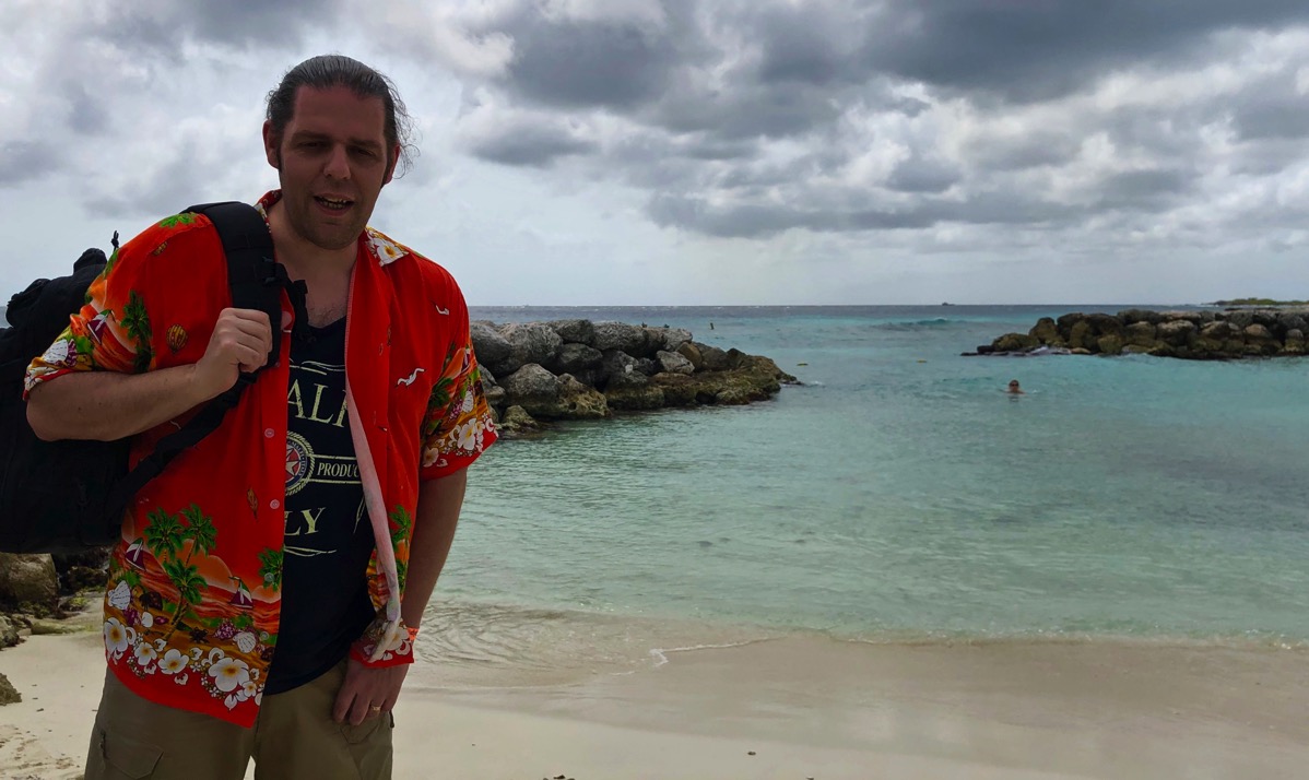 Me stood on the left in a orange caribbean shirt carrying a black backpack with views of palm island, Aruba in the background