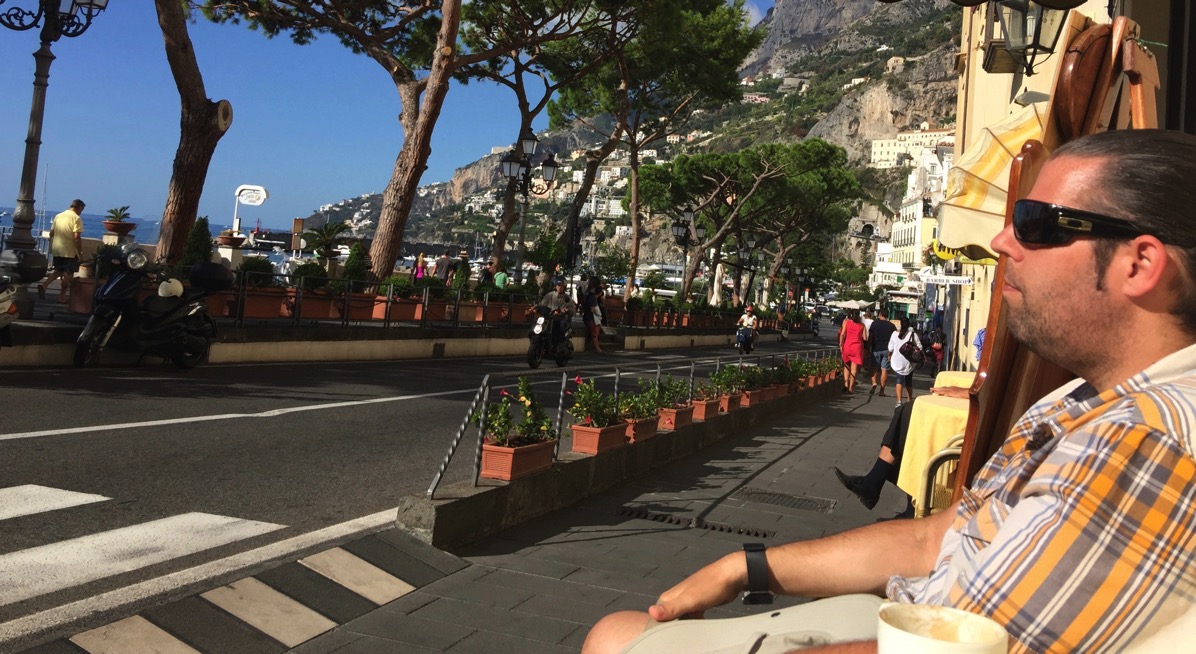 Me sat in the sun outside the cafe eating a coissant and drinking an espresso ith the road right in front of me