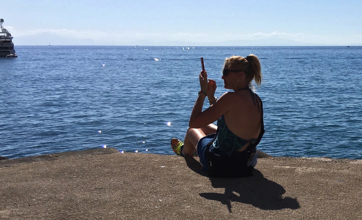 Sarah sat on the very small dock with her feet off the end taking a photo of a ship that is fust in shot