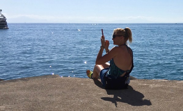 Sarah sat on the very small dock with her feet off the end taking a photo of a ship that is fust in shot