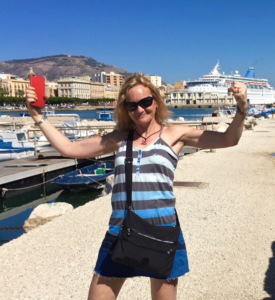 Sarah showing her guns in a victory pose stood in front of lots of colourful boats in the dock at Sicily