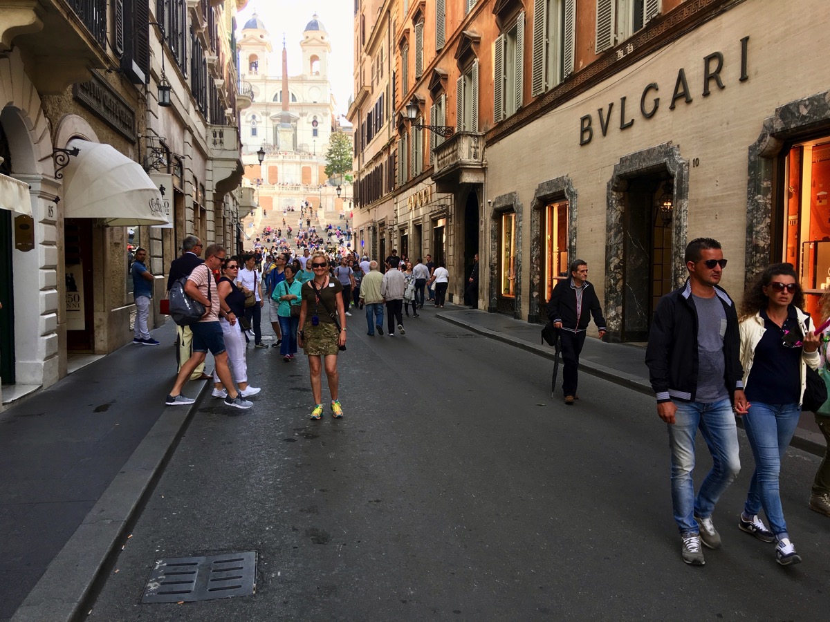 Sarah walking towards the camera down the centre of a busy street in Rome with tall building s on either side
