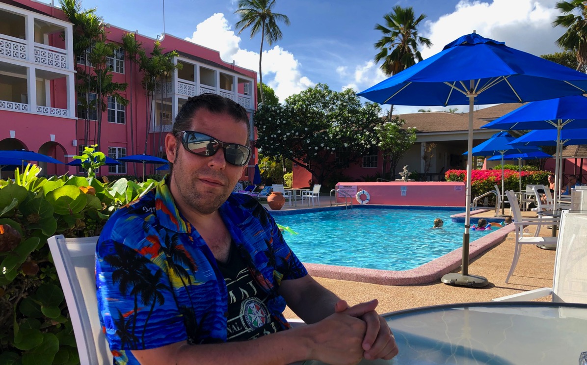 Me sat in blue Caribbean shirt next to the swimming pool and the beach