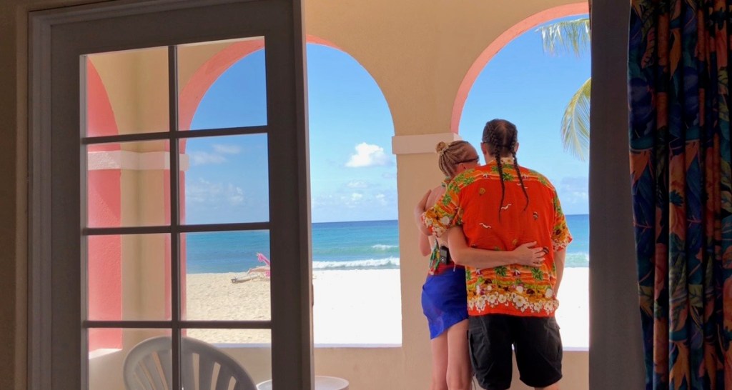 A couple embraces, gazing at the beach and ocean through an arched window, enjoying a romantic moment together.