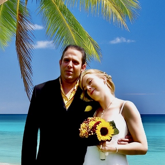 A couple stands together on a beach, embracing each other lovingly. The woman holds a bouquet of sunflowers and leans her head against the man's shoulder, surrounded by palm trees and a serene ocean backdrop.