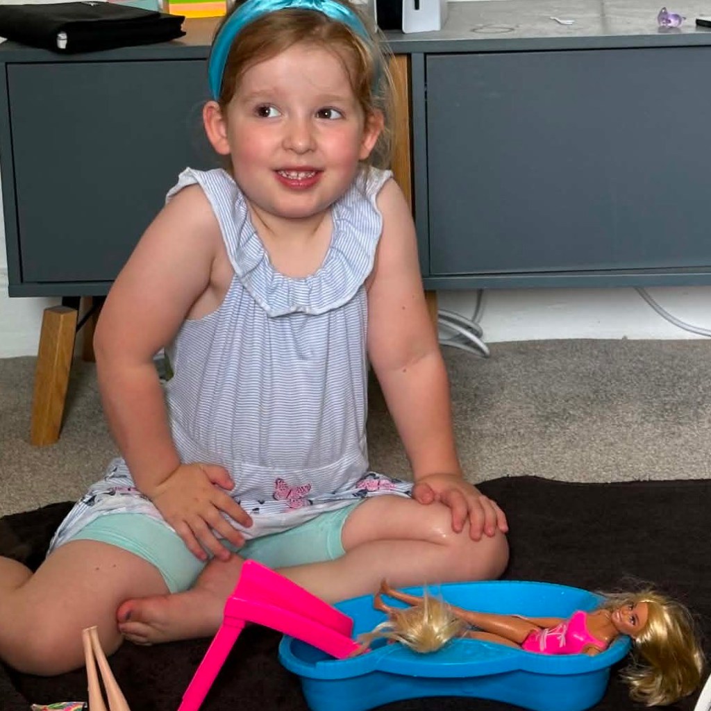A young girl, Jess, sitting cross-legged on a dark mat on the floor indoors. She is wearing a light blue and white striped sleeveless top with ruffled shoulders, light turquoise shorts, and a bright blue headband in her strawberry-blonde hair. She looks off to the side with a slight smile while playing with a blue toy pool set and fashion dolls in front of a grey side cabinet.