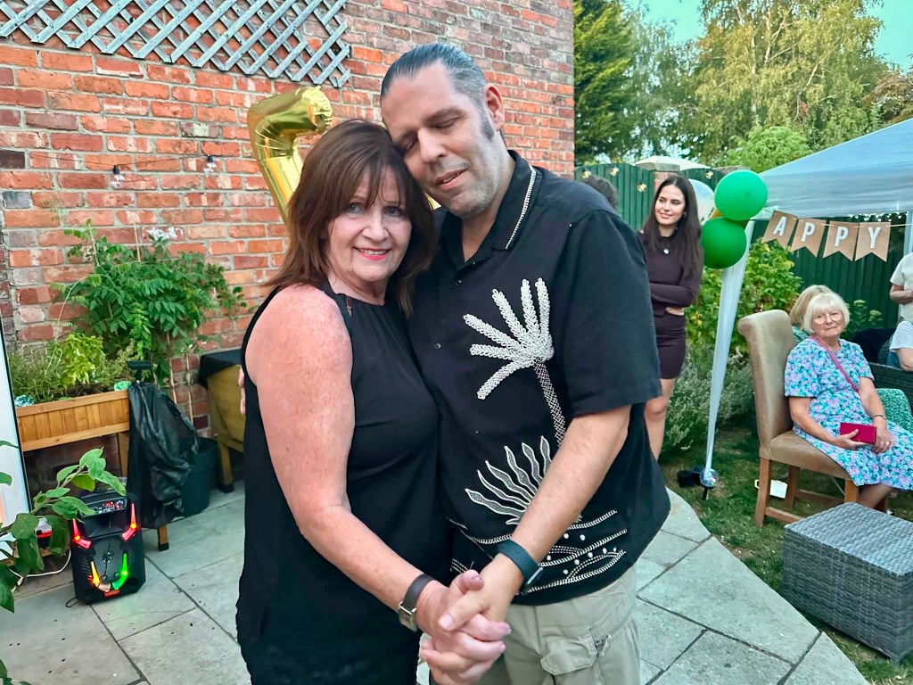 Bridge on the left, a woman with dark shoulder-length hair wearing a black sleeveless top, smiling as she holds hands and slow dances with Steve on the right, who has his eyes closed and his head resting gently against hers. They are outdoors in a paved patio garden during a celebration, with party decorations, green balloons, a lit-up party speaker playing music nearby, and family and friends gathering around them in the background.