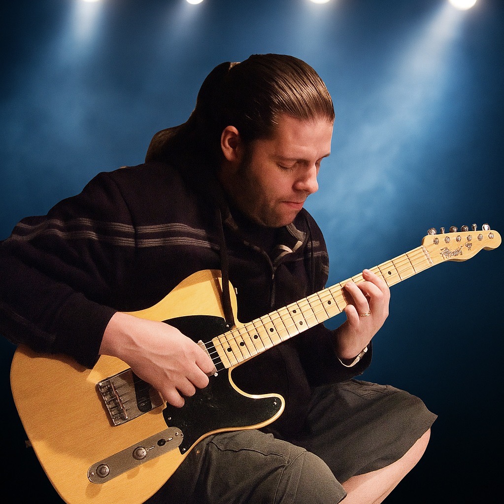You are seated and captured mid-performance, looking down with focused concentration at the fretboard.
You have long brown hair slicked back into a low ponytail, light facial hair, and are wearing a dark hooded sweater with subtle horizontal striped detailing across the chest and arms.
Instrument
You are holding a Butterscotch Blonde Telecaster featuring a classic single-cutaway ash body with visible wood grain under the warm finish.
The guitar is fitted with a black pickguard, a chrome control plate with volume/tone knobs and a pickup switch, a vintage-style bridge with barrel saddles, and a maple neck and fretboard with dot inlays.
Lighting & Background
The background features a dramatic, hazy deep blue backdrop lit from above by soft, bright overhead spotlights, creating an atmospheric stage lighting effect.