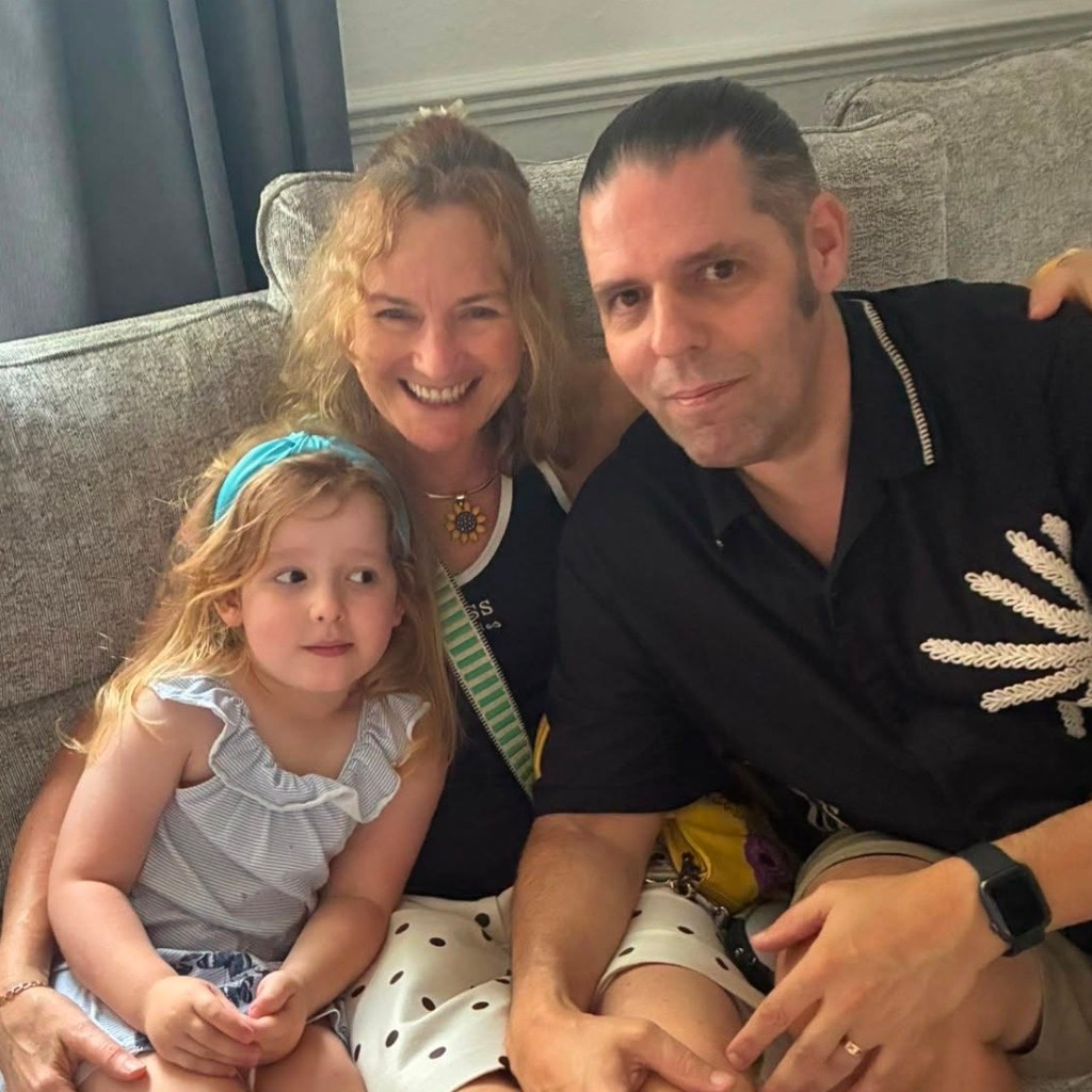Three people sitting closely together on a grey sofa indoors. On the left, Jess, a young girl wearing a light blue ruffled top and a blue headband, looks off to the side. In the middle, Sarah smiles warmly at the camera, wearing a dark top, a sunflower pendant necklace, and polka-dot bottoms. On the right, Steve sits beside her wearing a dark short-sleeved button-up shirt with an embroidered design, looking toward the camera.