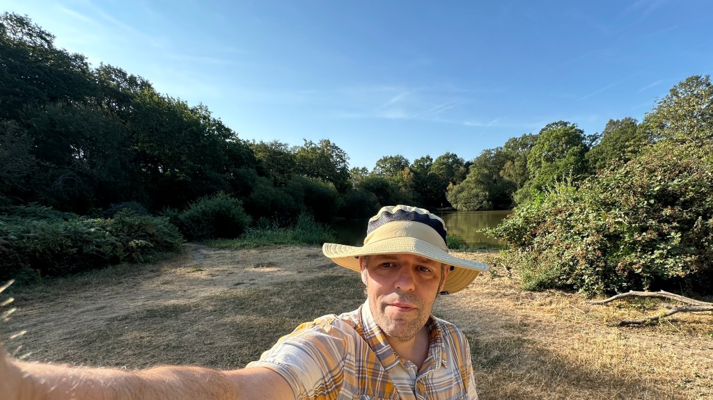 A self-taken photo on a bright, sunny morning by a tranquil pond. In the lower right foreground, Iβm wearing a wide-brimmed tan sun hat with a dark band and a yellow, white, and grey short-sleeved plaid shirt. Iβm holding the camera with my right arm extended. My expression is calm and content, with a gentle, relaxed half-smile and dark brown eyes looking directly toward the lens. The morning sun illuminates my face naturally under the shade of the brim. Behind me, the landscape opens up to a calm body of water surrounded by lush, dense green trees, wild bushes, and dry, sun-bathed grass under a clear blue summer sky.