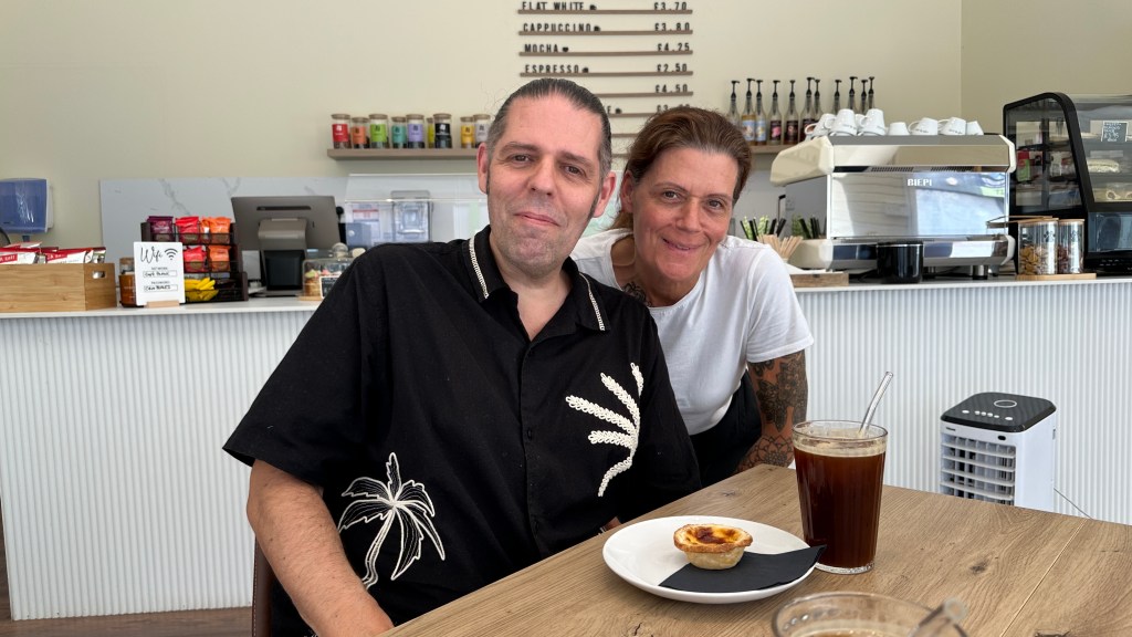 Steve on the left, wearing a black short-sleeved button-up shirt with white palm-like embroidered designs, seated at a wooden table with an iced coffee and a small pastry on a plate. Standing next to him on the right is his sister, Amber, wearing a white t-shirt, smiling warm-heartedly. They are inside her brightly lit coffee shop, with a white counter, espresso machine, menu board, and drink supplies visible in the background.