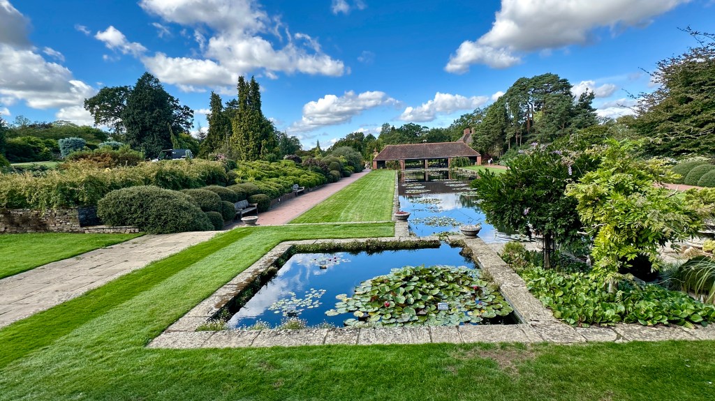 Alt Text: A wide-angle view of a formal garden featuring a square stone lily pond in the foreground reflecting a bright blue sky with fluffy white clouds. In the background, a long canal-style reflection pool stretches toward a brick pavilion, surrounded by manicured lawns, stone pathways, and lush green hedges.