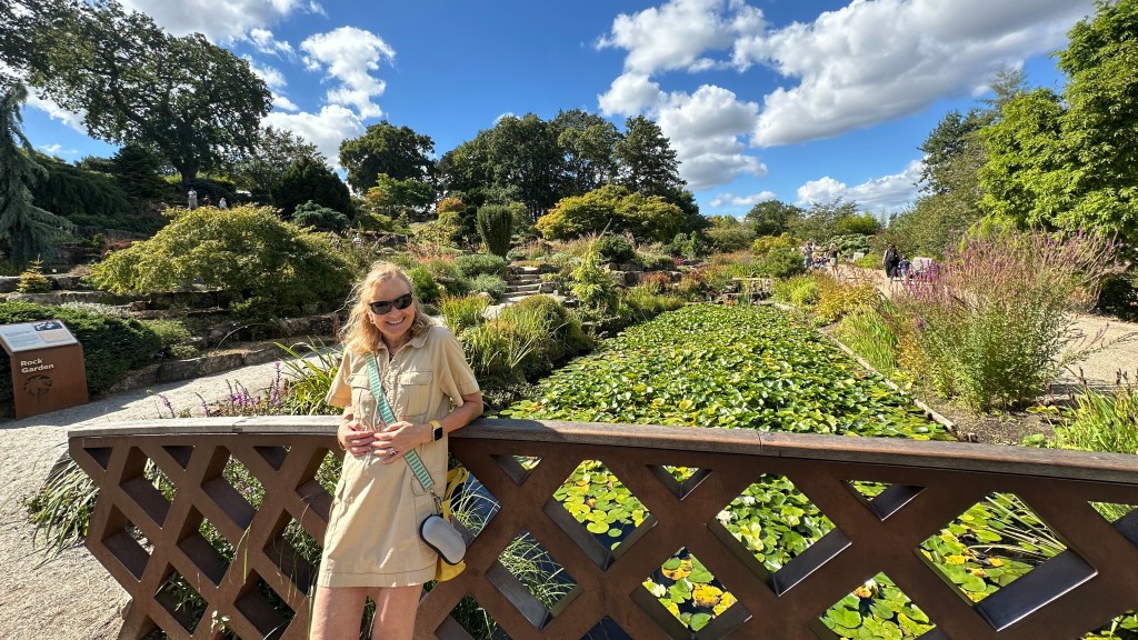 A blonde bombshell called Sarah smiling in sunglasses and a beige utility dress stands on a wooden wooden-latticed bridge. Behind her lies a long pond filled with green lily pads, flanked by lush botanical garden landscaping under a bright blue sky with fluffy white clouds. A sign reading "Rock Garden" sits on the left path.