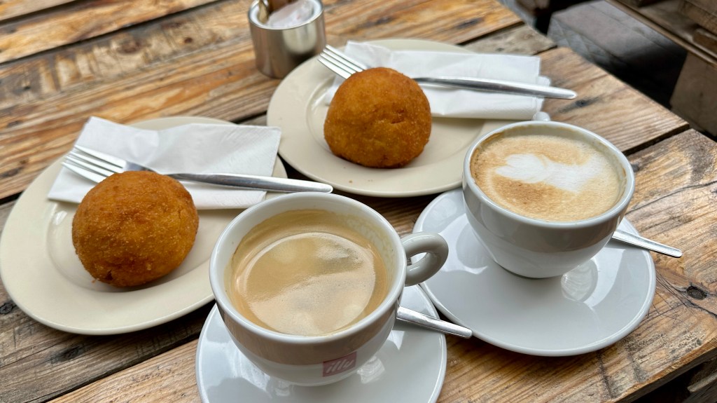 Detailed: A rustic wooden tabletop set with two white cups of flat white coffee with foam art, paired with two small plates holding round, fried arancini, forks, and white napkins.