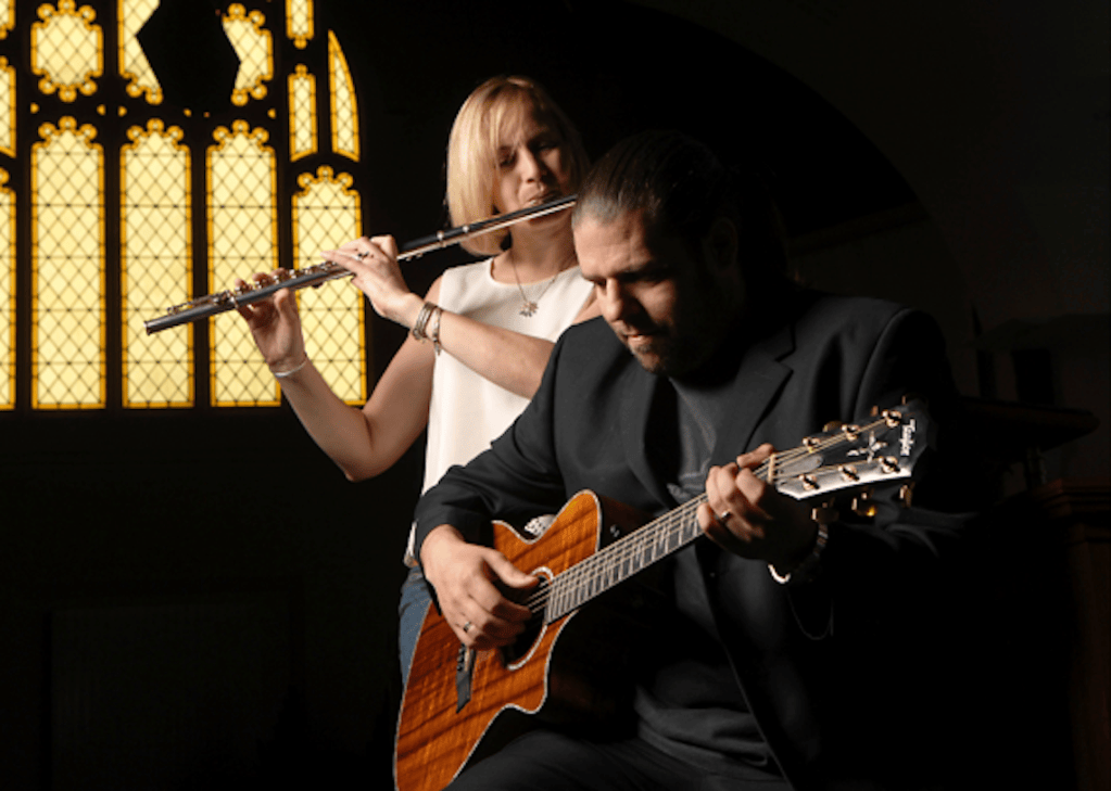 Image 6
An intimate, dimly lit photo inside a church featuring Steve and Sarah performing music together in front of a glowing stained-glass window. In the foreground, Steve sits angled slightly to the right, wearing a black blazer and t-shirt, focused on playing an acoustic Taylor K22ce guitar with a distinctive figure-grained koa finish. Standing just behind him to the left is Sarah, wearing a sleeveless white top and necklace, holding a silver flute horizontally and playing it with concentration. In the dark background, a tall, arched stained-glass window glows with warm yellow and amber light, creating a dramatic, ambient contrast behind the musicians.