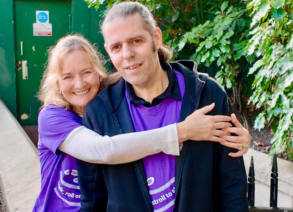Sarah smiling warmly while hugging Steve from behind with her arm wrapped around his shoulders, both standing close outdoors in front of lush green bushes. They are wearing matching bright purple MS Walk t-shirts featuring the event logo, with Steve wearing a dark hoodie over his shirt.