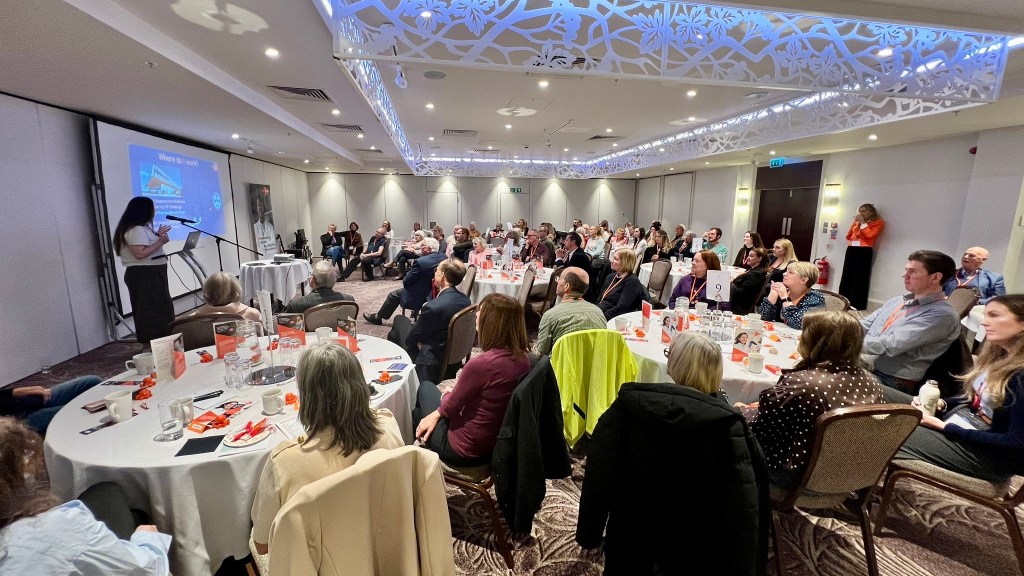 Guests seated around round tables in a conference room at an MS Society legacy event, listening to a speaker presenting information on a large screen at the front of the room. The tables are laid with refreshments, glasses, booklets and orange event materials, while attendees listen attentively.
