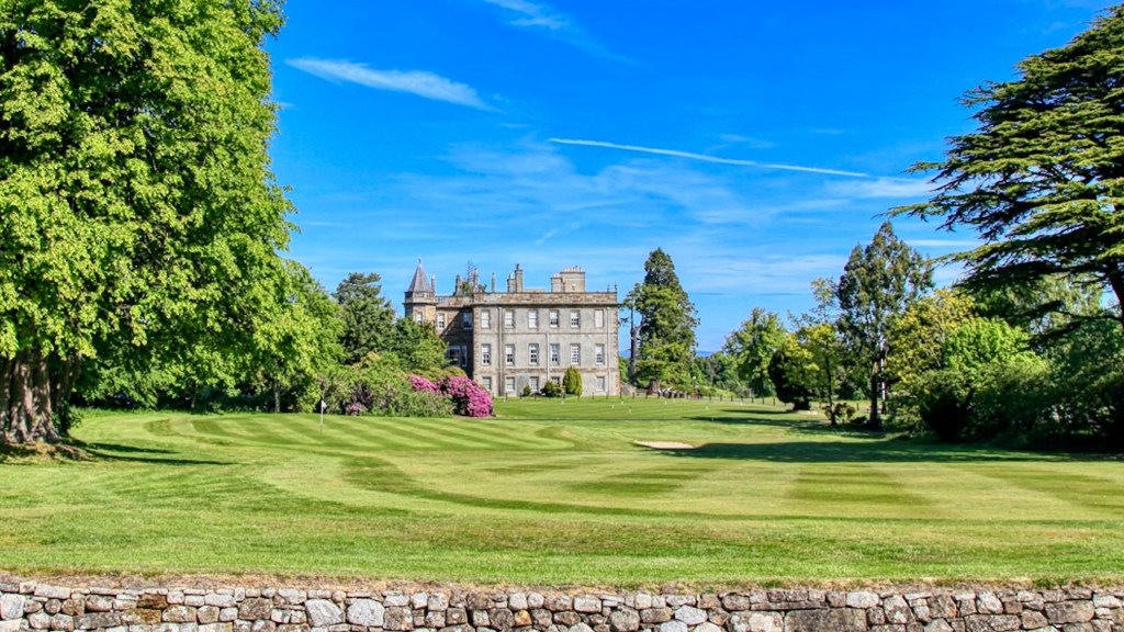 Dalmahoy House, a historic stone mansion set among beautifully landscaped green lawns and mature trees, with colourful rhododendrons, a stone wall in the foreground and a bright blue sky above.