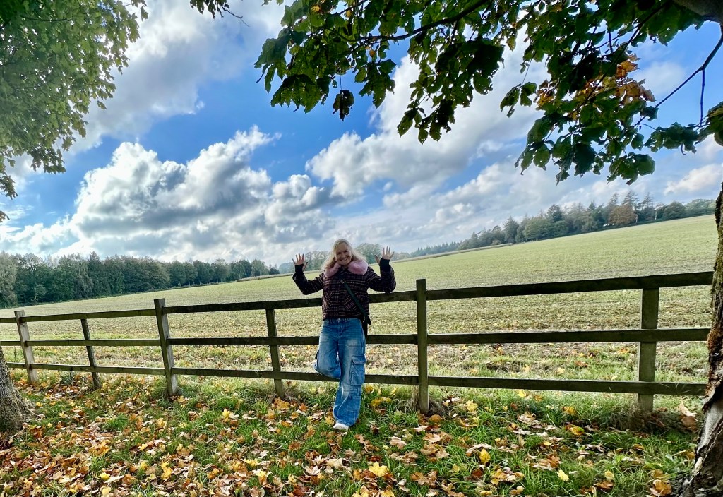 Sarah leaning against a wooden fence in a wide grassy field framed by large leafy trees under a bright blue, cloudy sky.   