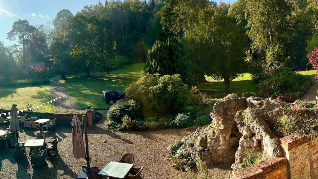 An outdoor patio area with tables and chairs next to a rock grotto formation and a lush, tree-lined lawn stretching into a forested hill.   