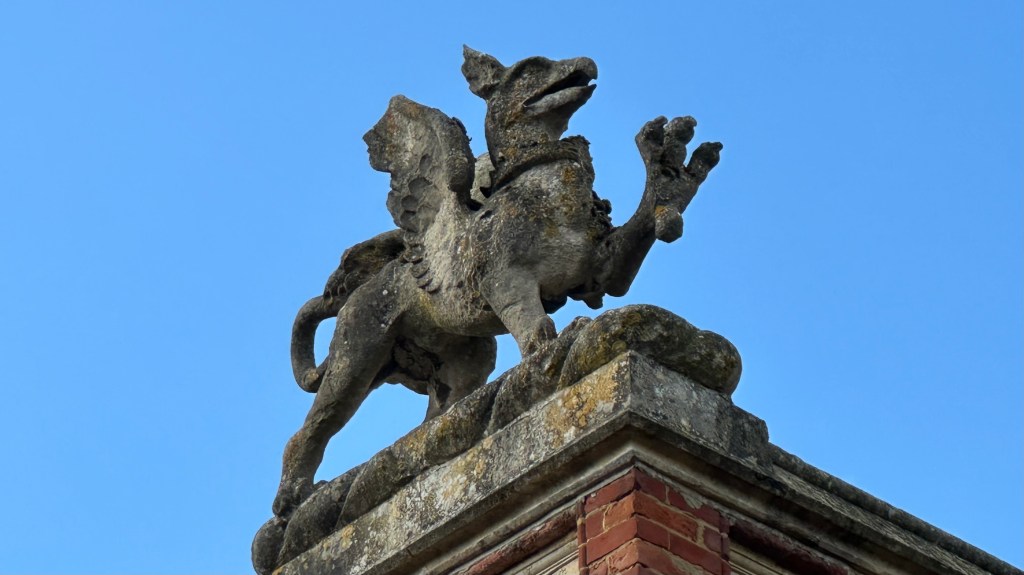 A close-up shot of a stone griffin statue standing atop the corner pillar of Wotton House against a clear blue sky.   