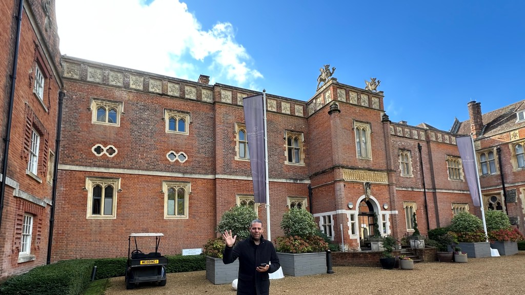 Steve (me) waving at the camera in front of a grand red-brick historic estate with gothic windows and decorative stone statues on top.   