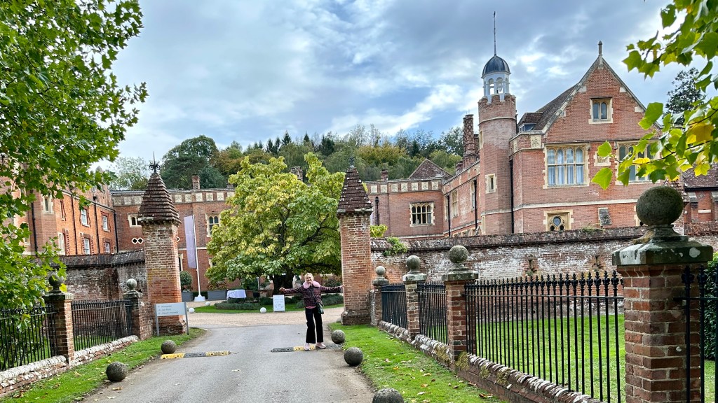 Sarah standing with arms outstretched on a paved driveway entrance to Wotton House with brick pillars and wrought iron fencing.   