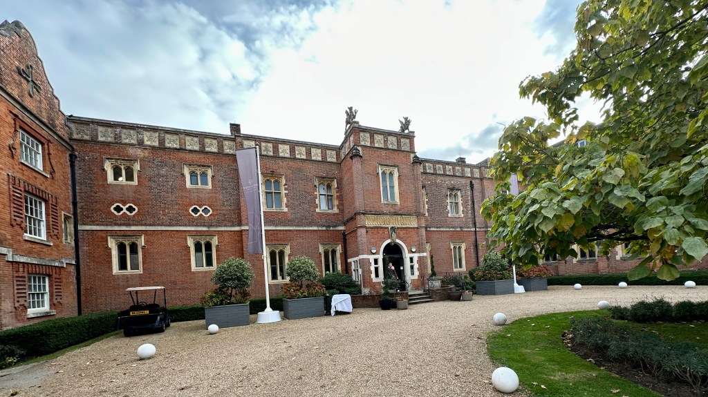 A wide courtyard paved with gravel leading to the main entrance of Wotton House with potted plants and green foliage. Sarah is stood at the entrance doors.