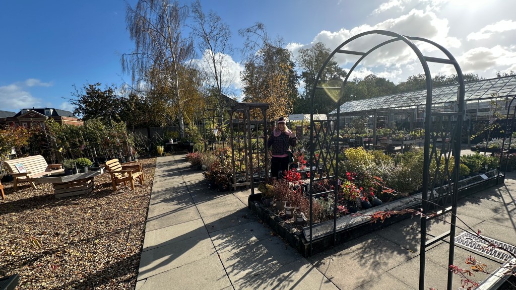 Alt Text: A wide outdoor shot of a garden plant nursery under a bright, partly cloudy blue sky. A paved concrete path runs through the center between gravel displays with potted plants, shrubs, wooden benches, and garden arches. A woman in a pink top, dark jacket, and beanie hat stands in the middle distance under a wooden archway, waving towards the camera. A large greenhouse and metal decorative arches are visible on the right.