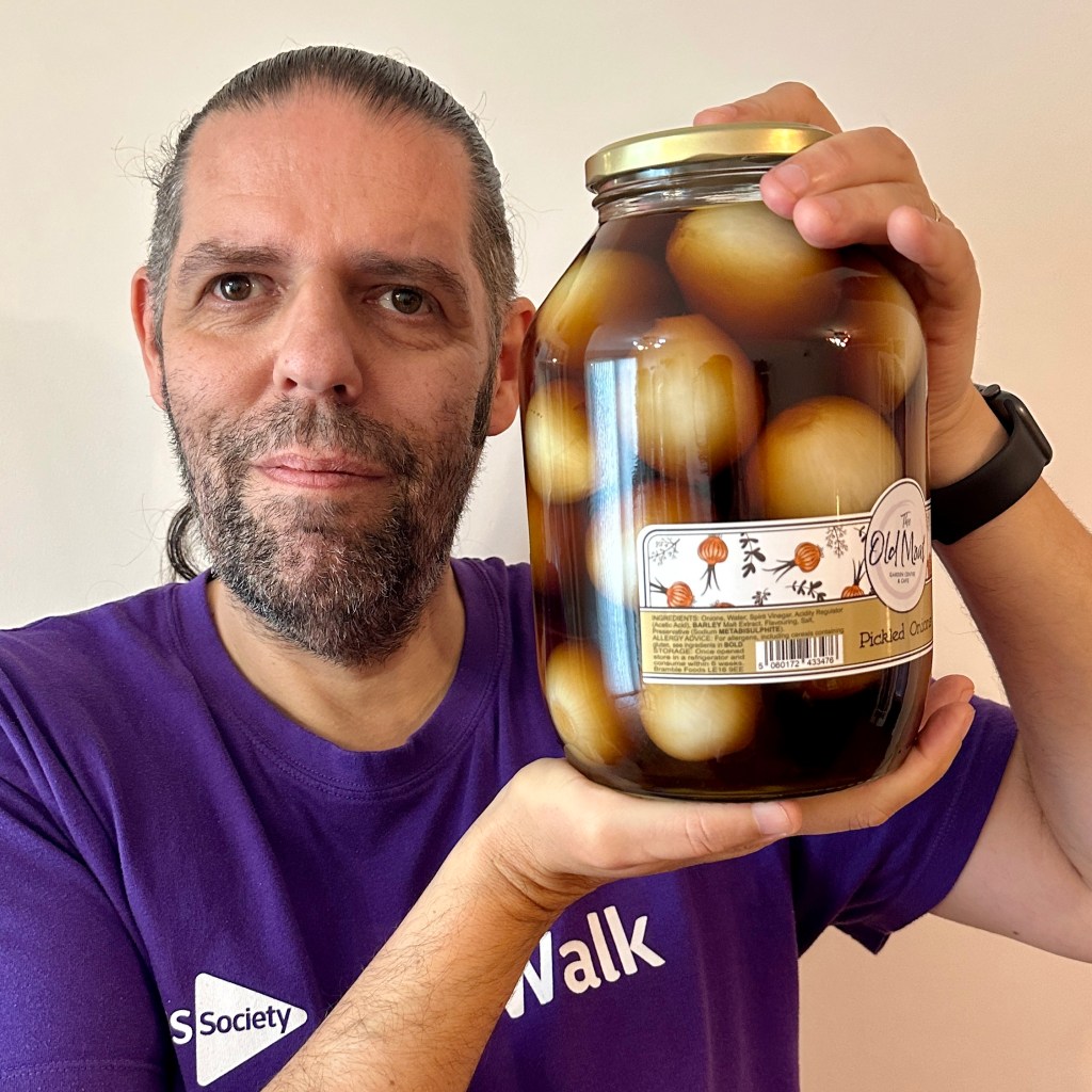 Alt Text: A close-up portrait of a man with light skin, short dark hair tied back in a small ponytail, and facial scruff, wearing a purple shirt that reads "MS Society Walk". He is smiling at the camera while holding up a large glass jar filled with pickled onions in dark vinegar. The jar features a label from "The Old Mill Garden Centre & Cafe".