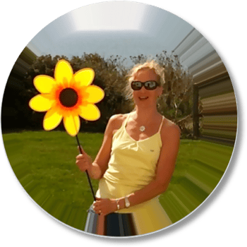 Sarah stands outdoors on a sunny day against a backdrop of a green lawn and dark bushes, wearing dark sunglasses, a necklace, and a yellow spaghetti-strap top. She is smiling while holding a large, bright yellow toy flower on a stick in her right hand. The entire photograph is set within a circular frame that features a distinct radial stretch and "sunburst" warping effect radiating outward from the center.   